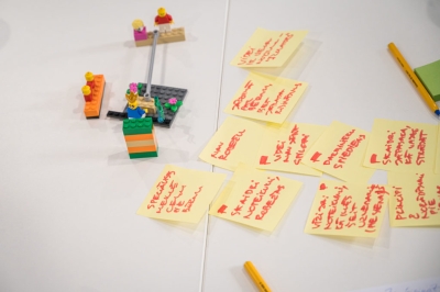 Līmlapiņas ar pierakstiem un neliels LEGO modelis uz darbnīcas galda.