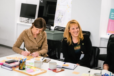 Divas darbnīcas dalībnieces sēž pie galda; viena pieraksta idejas, otra klausās diskusiju.