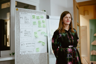 Dalībniece stāv pie flipčarta ar uzrakstiem "Problēmas" un  numurētiem jautājumiem par veidlapām un valodu, ap kuriem piekārtotas zaļas piezīmju lapiņas.